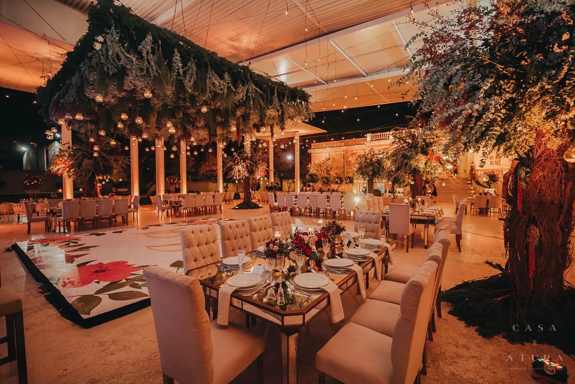 Banquete de boda en los jardines con pista floral, mesas espejo, candelabros y follaje suspendido con luces